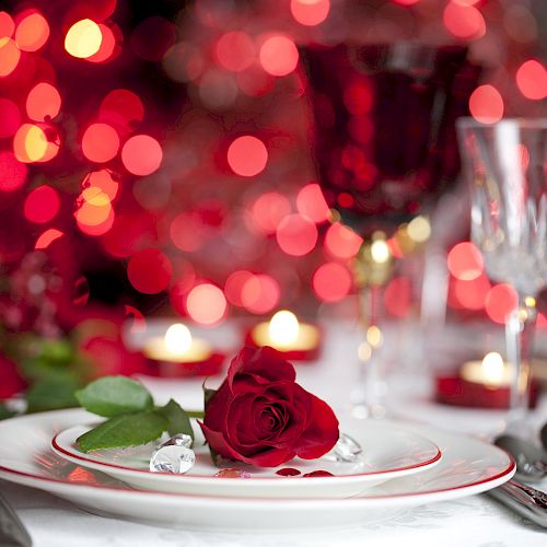 A romantic dinner setting with a red rose on a plate, surrounded by red lights, candles, and glasses in the background.