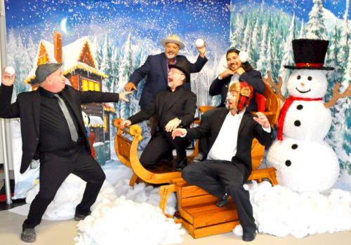 Five people are playfully posing in a winter-themed setting with a sleigh, snowman, and snowballs, in front of a snowy backdrop.