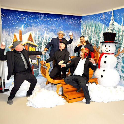 Five people are playfully posing in a winter-themed setting with a sleigh, snowman, and snowballs, in front of a snowy backdrop.