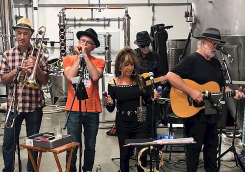 A band of five musicians, including a guitarist, singer, harmonica player, and trombone player, performing in a distillery setting with equipment around.