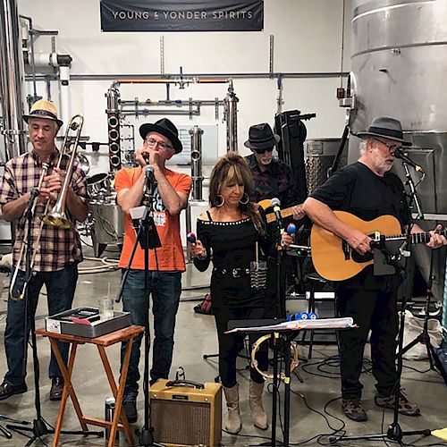 A band of five musicians, including a guitarist, singer, harmonica player, and trombone player, performing in a distillery setting with equipment around.