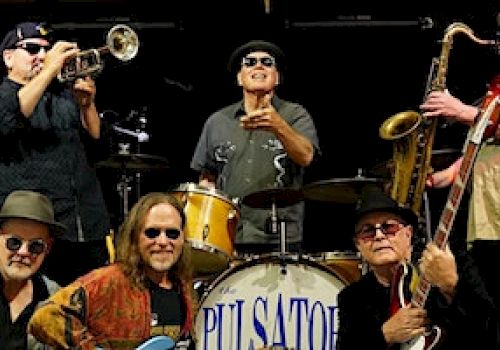 The image shows six musicians with various instruments like trumpet, saxophone, drums, and guitars, posing together in front of a drum set.