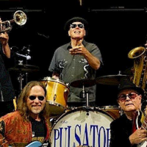 The image shows six musicians with various instruments like trumpet, saxophone, drums, and guitars, posing together in front of a drum set.