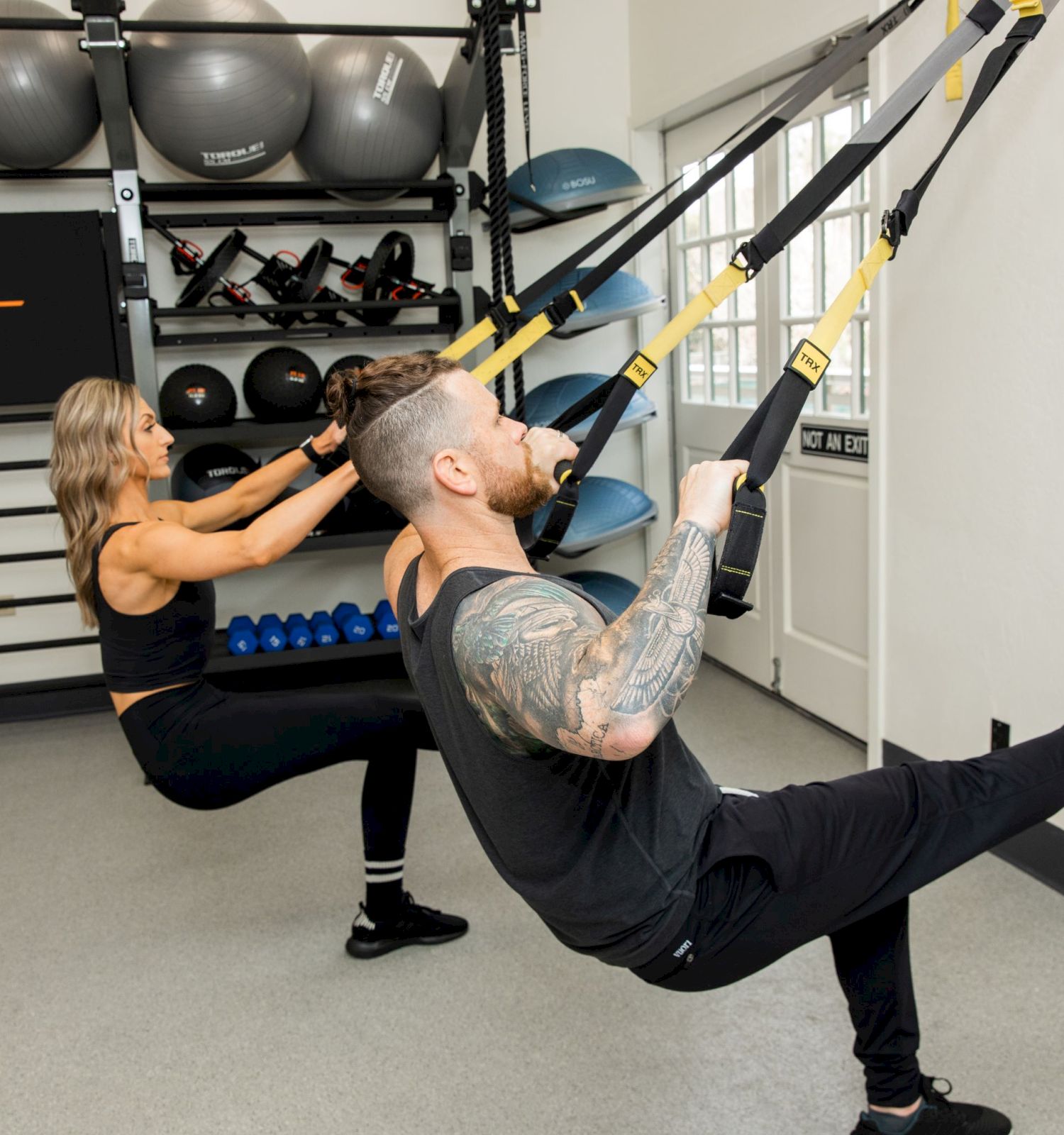 Two people are using TRX suspension training bands attached to a wall in a fitness room, exercising with one leg off the ground.