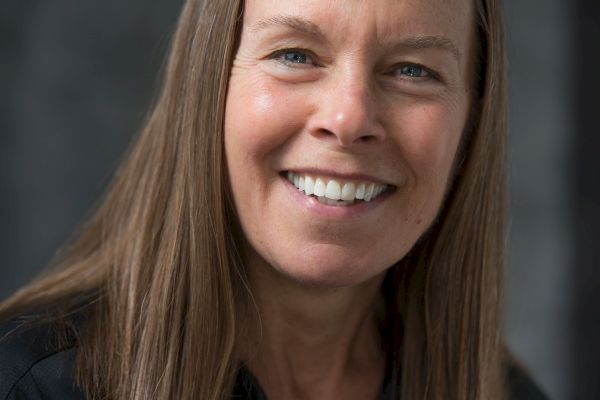 A smiling woman with straight, shoulder-length hair is wearing a dark shirt. She is standing against a dark, neutral background.