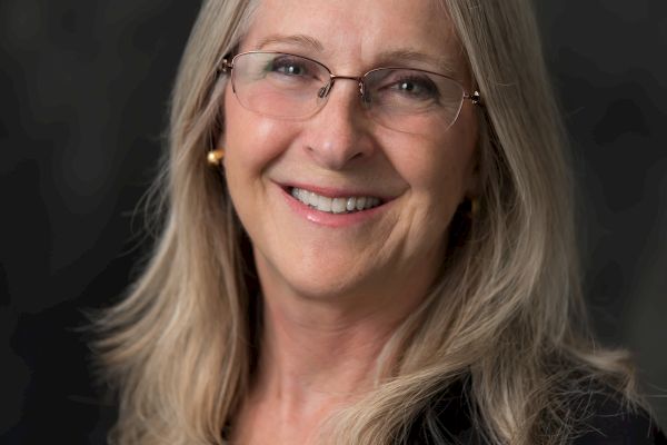 A person with long, blonde hair and glasses is smiling, wearing a black shirt with an embroidered logo. The background is dark.