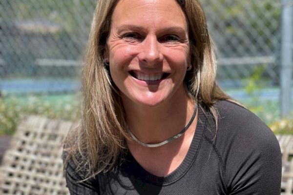 A person with long blonde hair is smiling and wearing a black top while sitting outside on a bench; there's a chain-link fence and greenery in the background.