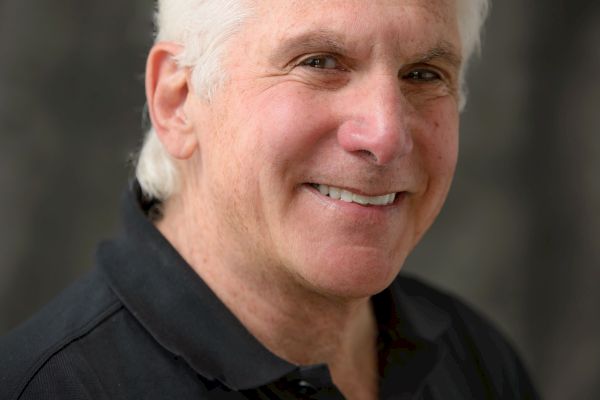 A man with white hair smiles at the camera, wearing a dark collared shirt with a subtle logo, set against a blurred background.