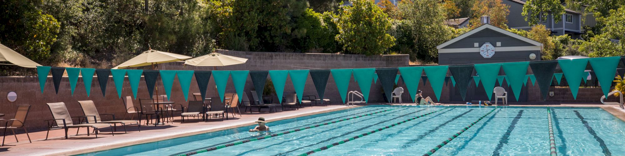 The image shows an outdoor swimming pool with multiple lanes, swimmers in the water, lounge chairs, umbrellas, and trees in the background.