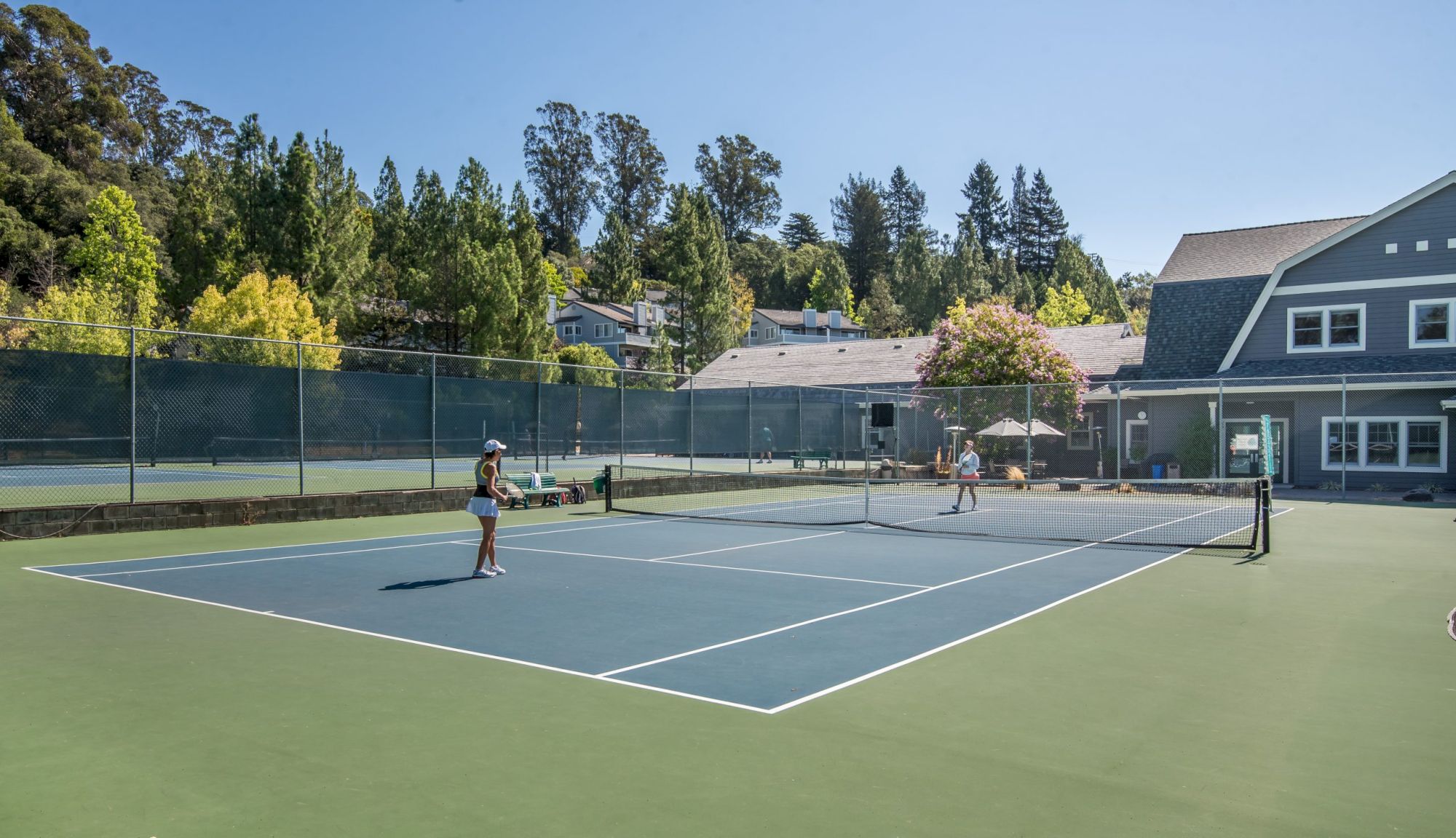 Two people are playing tennis on an outdoor court surrounded by trees and buildings.