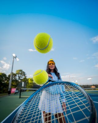 A person on a tennis court is hitting two tennis balls with a racket, with a blue sky in the background.