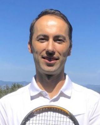 A man in a white shirt is standing outside, holding a tennis racquet. The background features a blue sky and distant mountains.