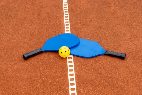 The image shows two blue pickleball paddles crossed over each other with a yellow perforated plastic ball placed on top, on a court.