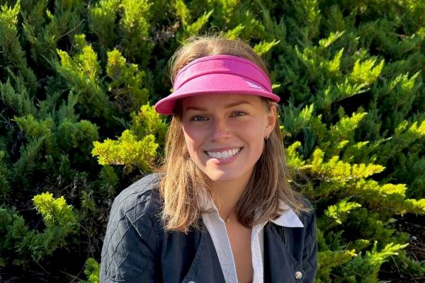 A person wearing a magenta visor and black jacket is smiling in front of lush green bushes and trees, enjoying a sunny day outdoors.