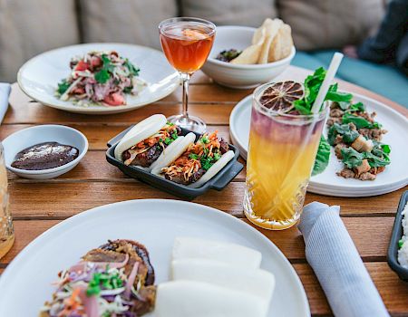 The image shows a variety of dishes and drinks on a wooden table, including tacos, salads, cocktails, and plates of food, all set for a meal.