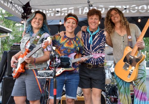 Four smiling people hold musical instruments under a canopy, suggesting a casual outdoor concert setting.