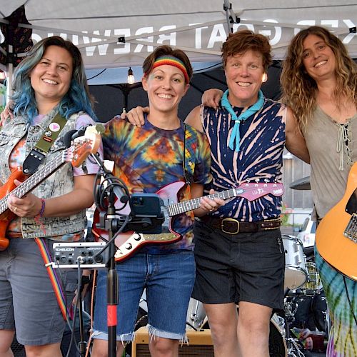 Four smiling people hold musical instruments under a canopy, suggesting a casual outdoor concert setting.