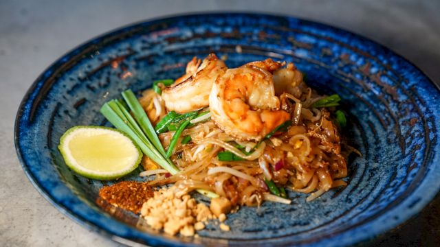 A plate of pad Thai with shrimp, lime wedge, peanuts, and green onions on a blue dish.