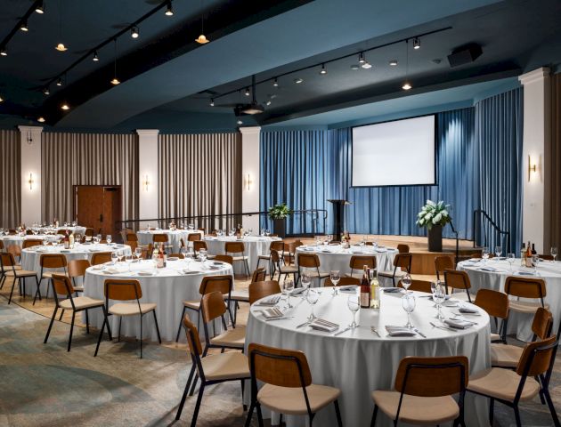 A banquet hall set up for an event, with round tables, chairs, table settings, wine bottles, and a stage with a projector and blue curtains ending the sentence.