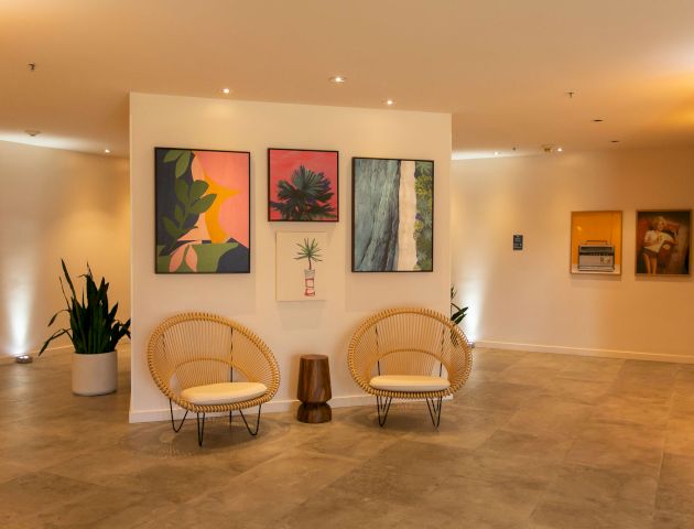 This image features a modern interior with two wicker chairs, a small wooden table, plant decor, and colorful framed artwork on the walls.