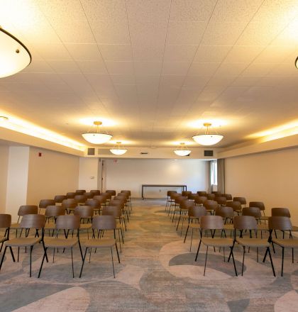 The image shows an empty conference room with rows of chairs arranged facing a table at the front, under ceiling lights, ready for a meeting or event.