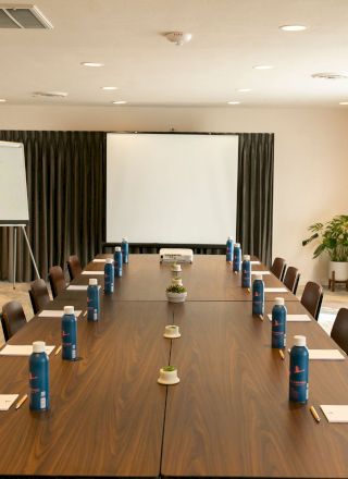 A conference room with a long table, chairs, water bottles, notepads, a whiteboard, a projector screen, and a plant by the window.