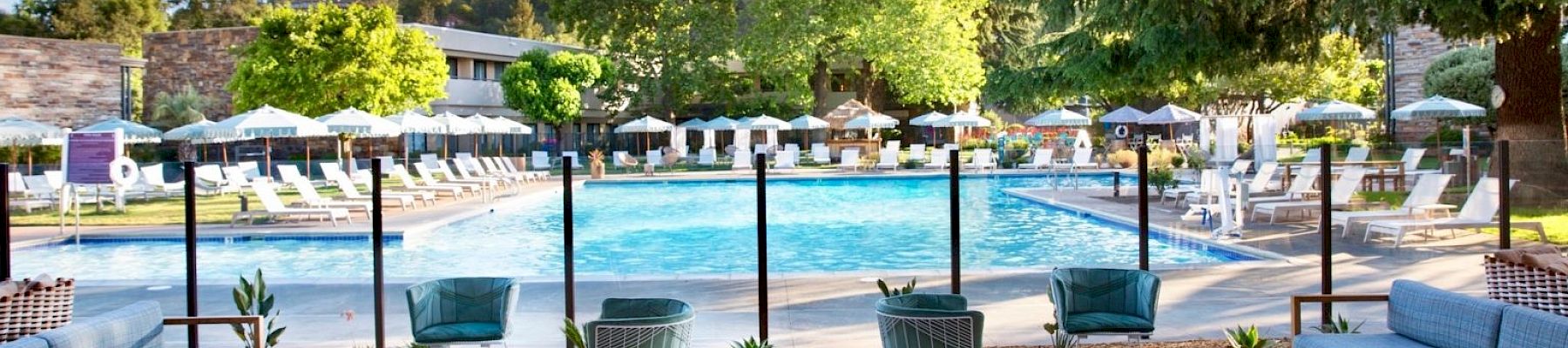 The image shows a pool area with lounge chairs, umbrellas, and seating arrangements under a wooden pergola, surrounded by greenery and buildings.