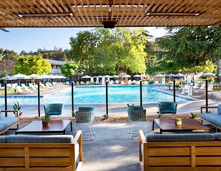 The image shows a pool area with lounge chairs, umbrellas, and seating arrangements under a wooden pergola, surrounded by greenery and buildings.