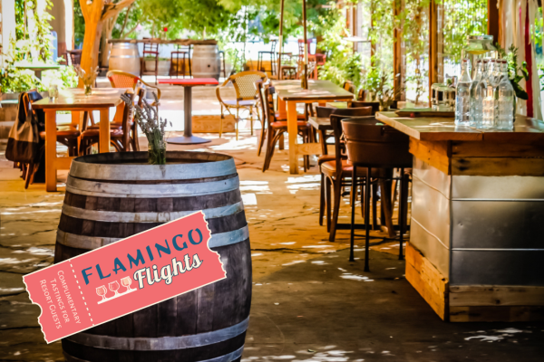 An outdoor seating area with wooden tables, chairs, barrel decor, and a bar. There's also a "FLAMINGO Flights" sign in the foreground.