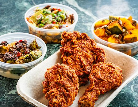 The image shows a plate of fried chicken drumsticks with three containers of various side dishes, including vegetables and salad.