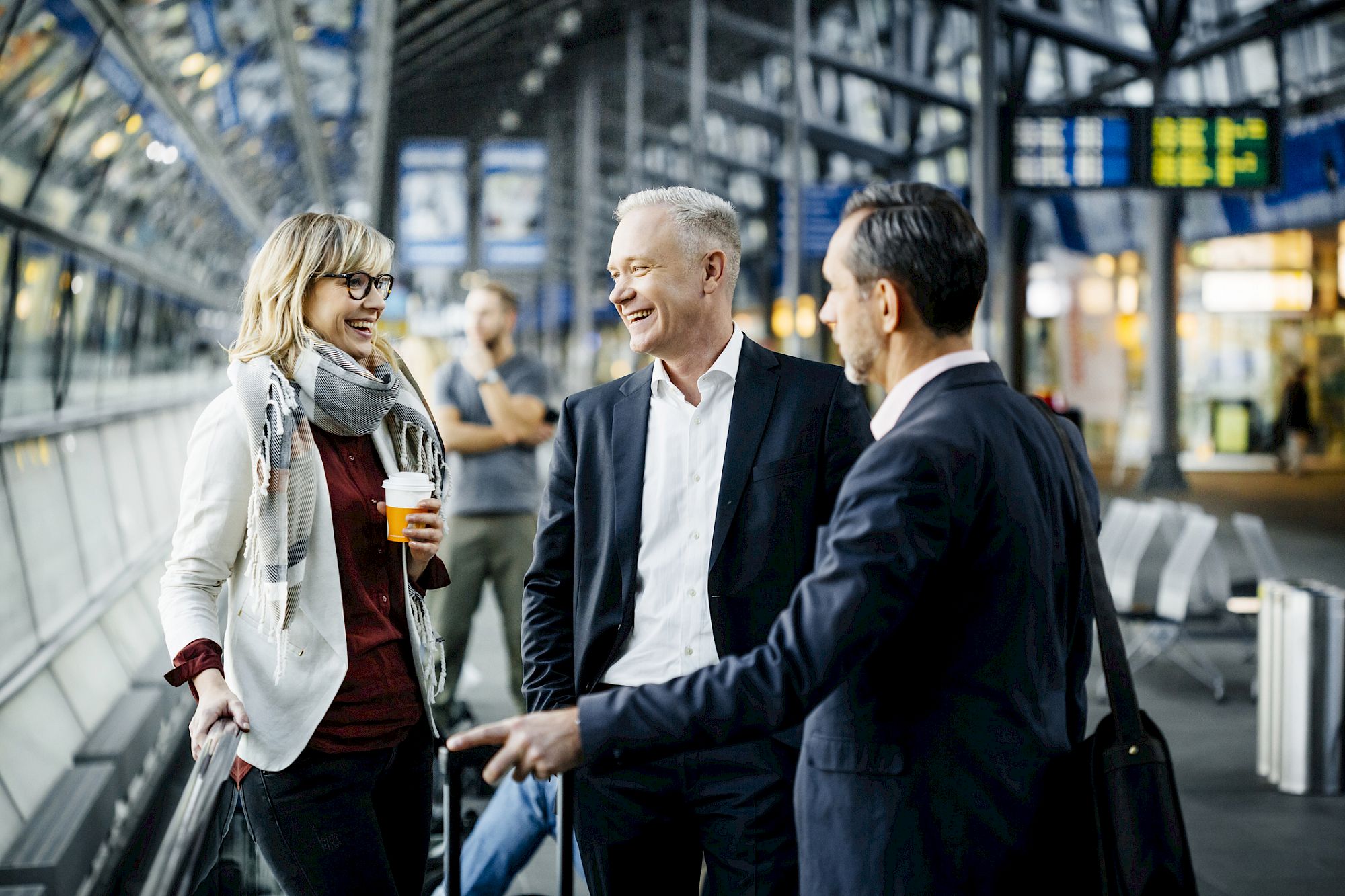 Three people are conversing in a modern, well-lit transport terminal, with one woman holding a drink and two men in suits, creating a lively atmosphere.