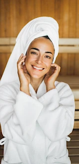 A person in a spa setting, wearing a white robe and towel on their head, smiling while seated on wooden benches.