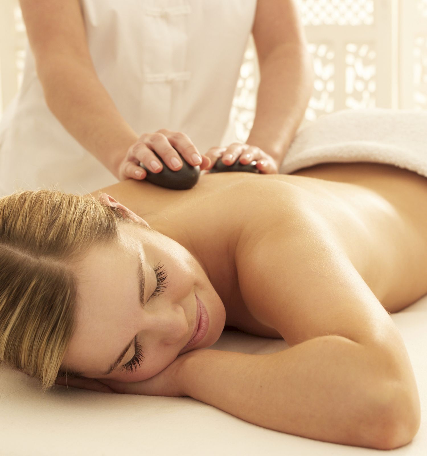 A person is receiving a back massage with hot stones, lying face down and covered by a towel, with a massage therapist attending to them.