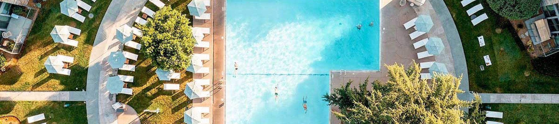 Aerial view of a rectangular swimming pool surrounded by lounge chairs, umbrellas, trees, and paths in a neatly landscaped area outdoors.