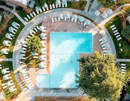 Aerial view of a rectangular swimming pool surrounded by lounge chairs, umbrellas, trees, and paths in a neatly landscaped area outdoors.