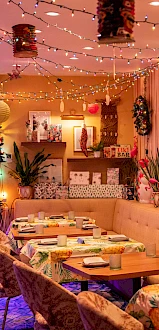 A cozy, colorful restaurant interior with decorative lights, floral patterns, and set tables, creating a festive and inviting atmosphere.