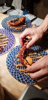 Hands plating an octopus dish with sauce on a patterned blue plate in a kitchen setting, with two more similar plates nearby.