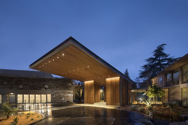 The image shows a modern architectural entrance with a wooden roof at night, featuring lighting and surrounded by landscaped areas.