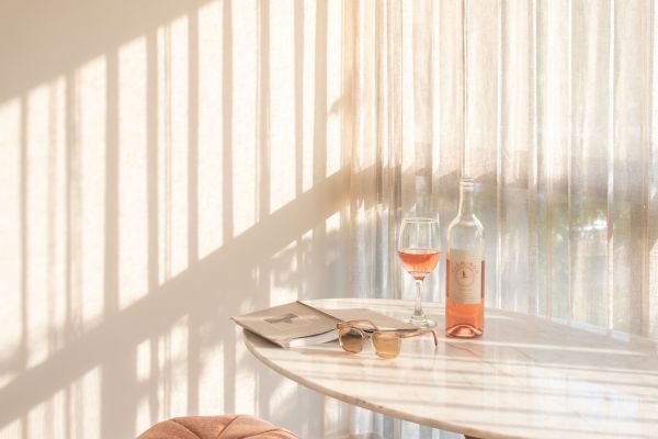 The image shows a cozy room with a round table, a pink stool, a bottle of rosé wine, and a filled glass near sheer curtains casting shadows.