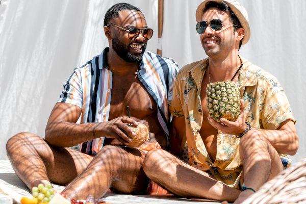 Two people are sitting and smiling in a sunlit, relaxed outdoor setting, each holding a drink, against a backdrop of white curtains.