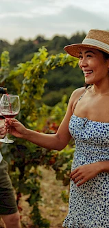 People enjoying wine while standing amongst lush vineyard rows, one pouring a drink for a smiling woman in a hat, others holding wine glasses.