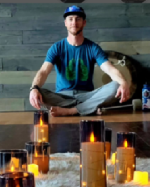 A person sits cross-legged on a wooden floor with a fuzzy rug and multiple lit candles in glass holders, creating a calming ambiance.