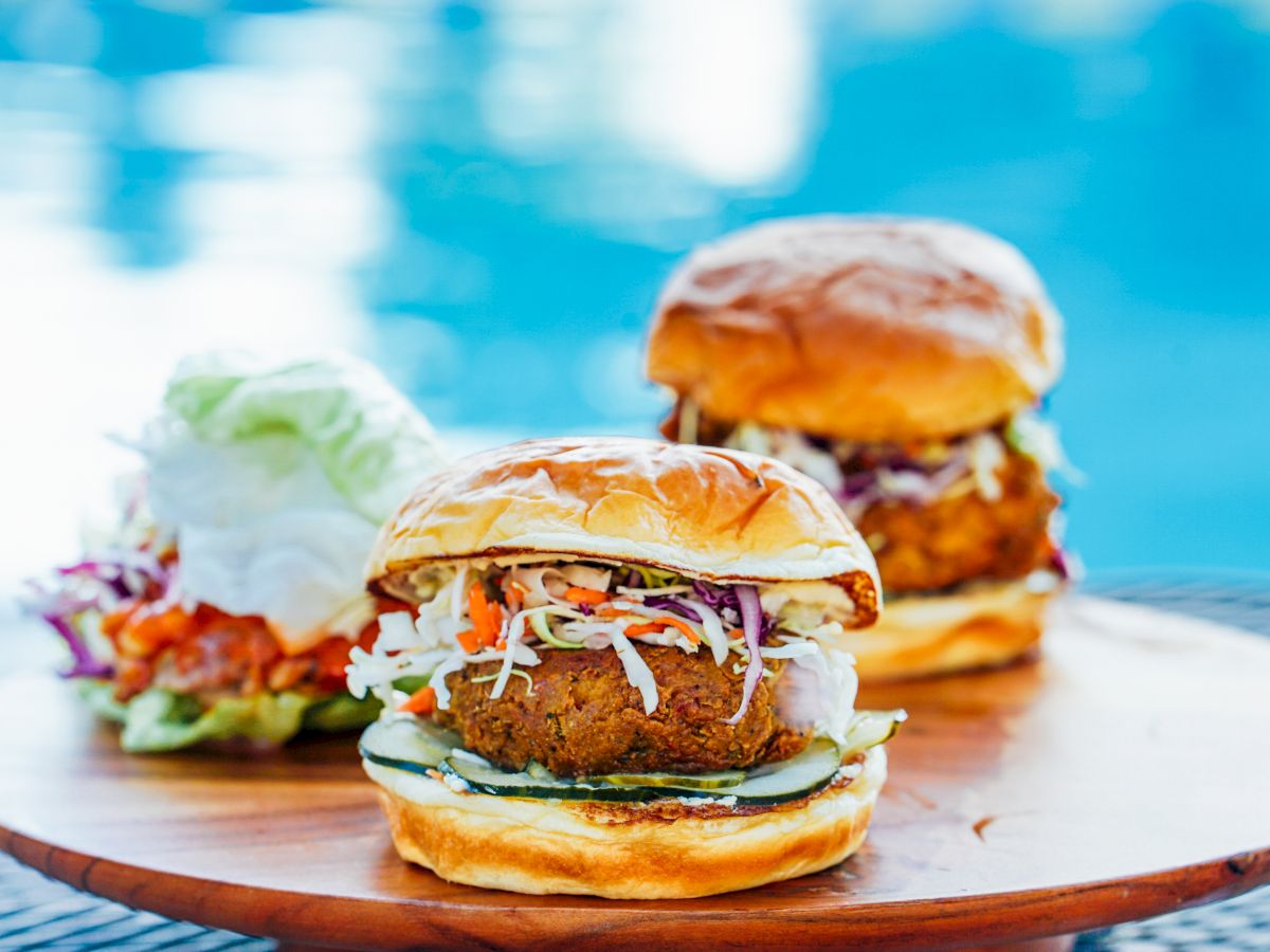 Two fried chicken sandwiches with coleslaw and cucumber on a wooden board, near a pool in the background, with a side salad.