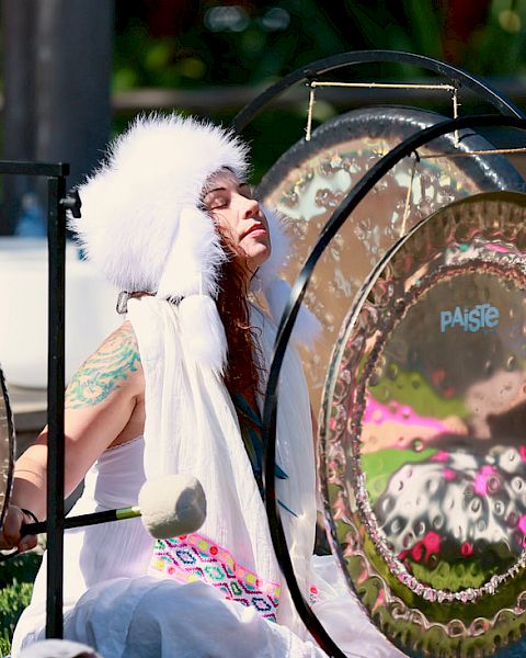 A person wearing a white headdress and white outfit plays large gongs outdoors, surrounded by greenery and sunlight.