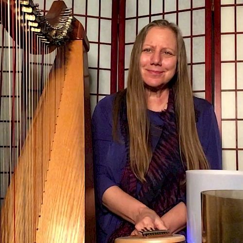 A person with long hair sits among musical instruments, including a harp and several other items, in front of a folding screen.