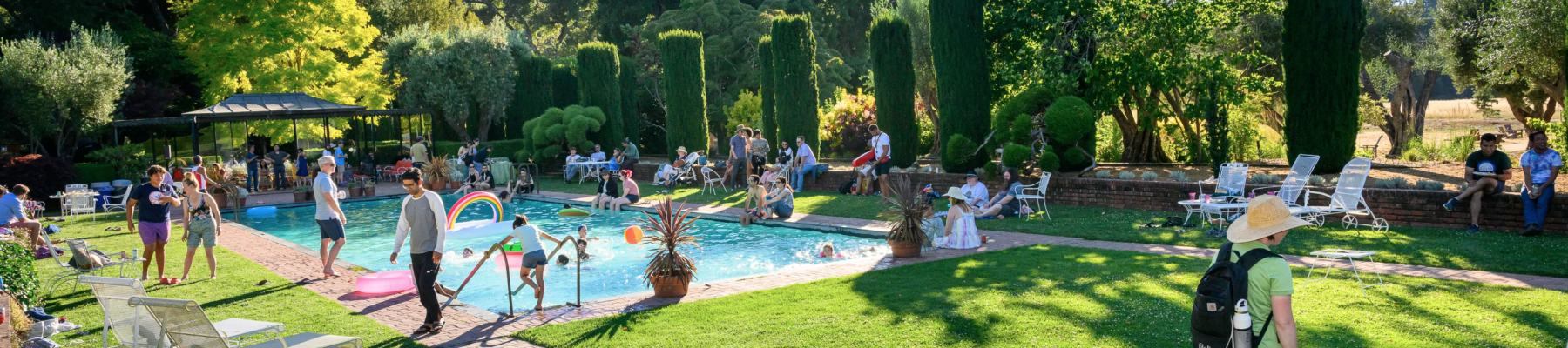 People enjoying a sunny day by the pool in a lush, green garden area with mountains in the background and various leisure activities happening.