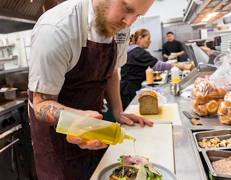 A chef in a kitchen is drizzling a yellow sauce onto a dish, with other staff and ingredients visible in the background.