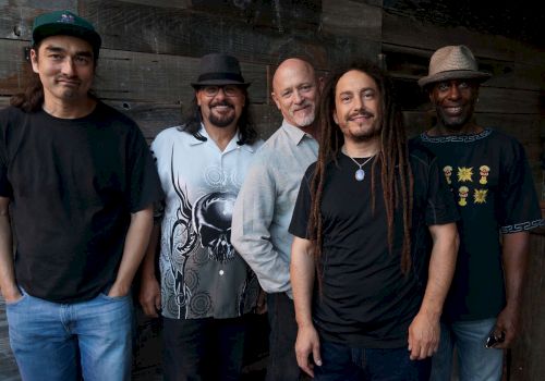 The image shows five men standing in front of a wooden wall, casually dressed, some wearing hats, all smiling at the camera, with various styles of clothing.