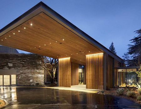 This image shows a modern building entrance with a large, illuminated wooden canopy and landscaped surroundings in the evening.