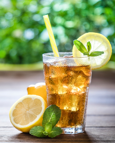 The image shows a refreshing iced tea with lemon slices and mint leaves on a wooden table, accompanied by a yellow straw, and a green, blurred background.